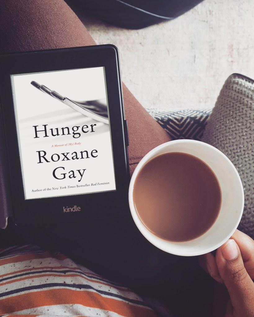 top view of a kindle on a lap with the cover of the book Hunger by Roxane Gay. Person on the sofa is holding a coffee cup next to it
