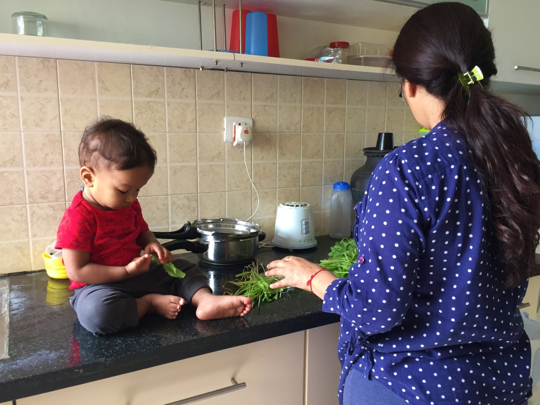 baby sitting on the kitchen counter with a spinach leaf while a woman cleans up a bunch of spinach