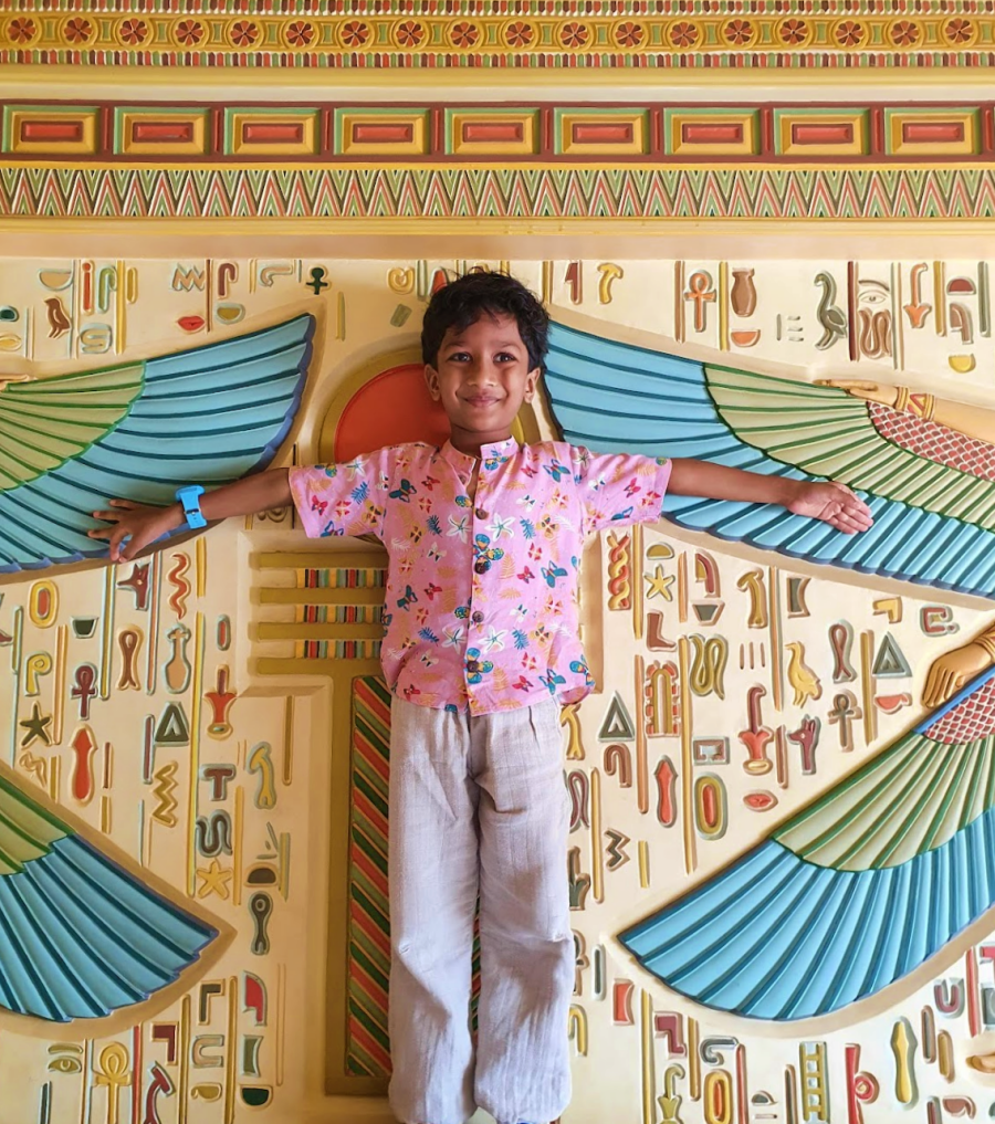 child in pink spreading his arms out in front of a colouful egyptian themed wall panel