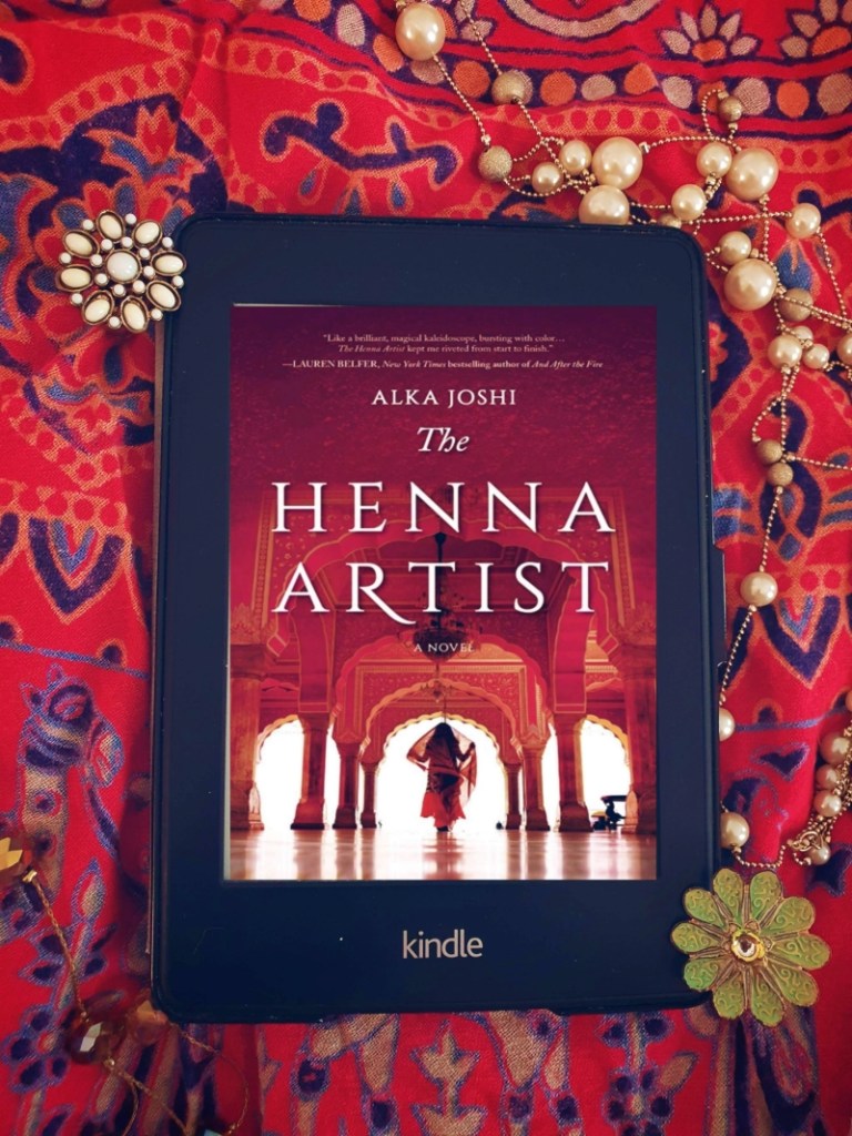 Kindle Paperwhite displaying cover of the henna artist laid on a bright red cloth with jewellery
