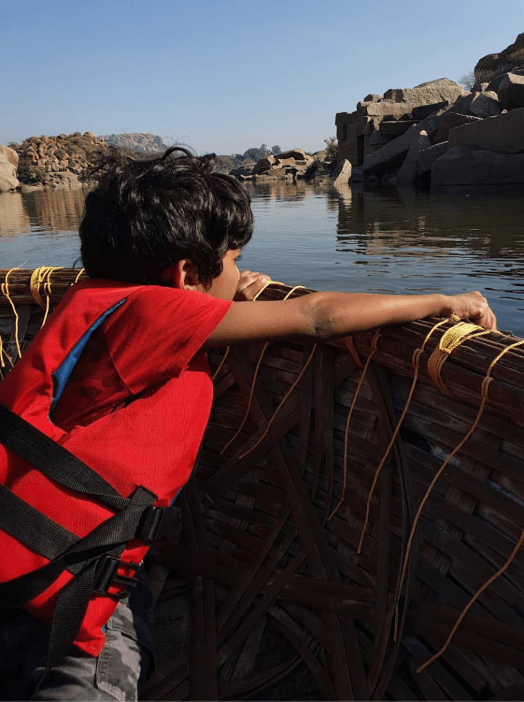 child in lifejacket looks over the coracle boat edge into the waters in hampi