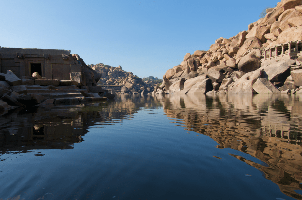 boulders and ancient temple ruins on either side of a small river reflected in the waters during the day while on a coracle ride in hampi