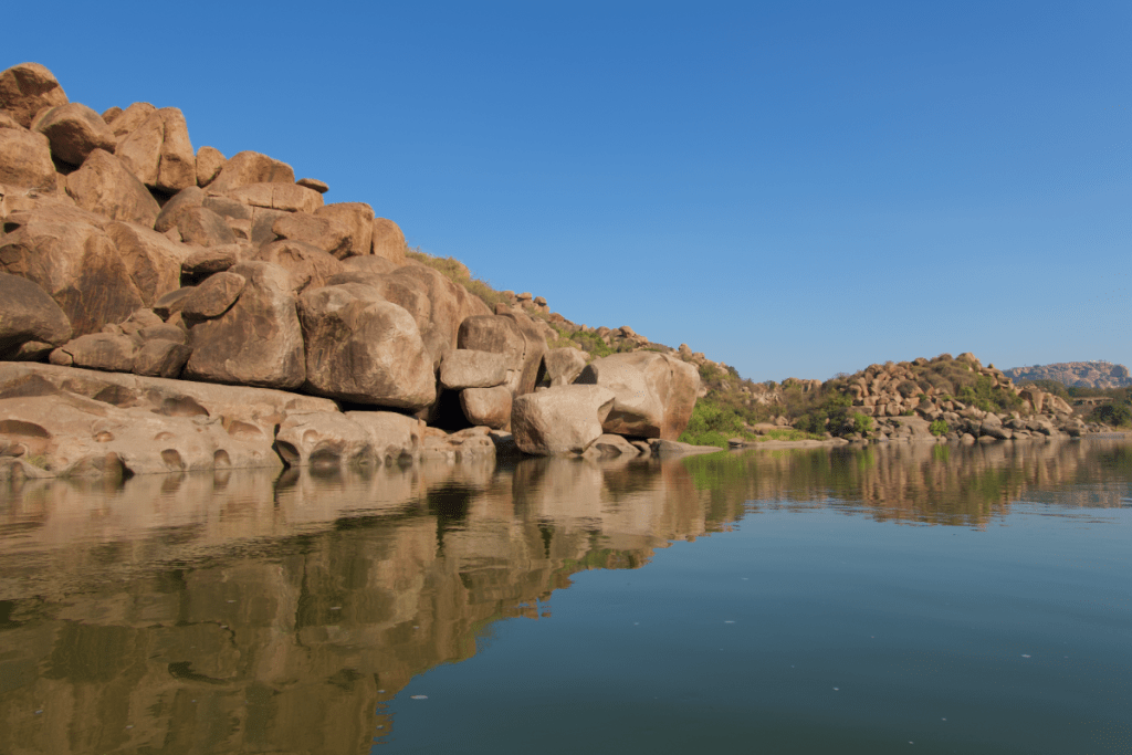 boulders line the side of a small river reflected in the waters during the day while on a coracle ride in hampi