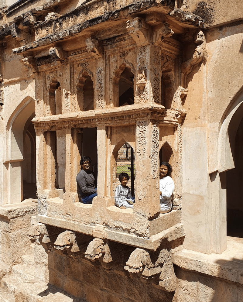 3 people looking out from one of the ornately carved stone window sit outs at Hampi Queen's Bath