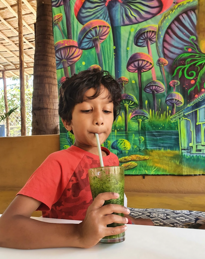 child in red shirt drinking a lemon and mint cooler with a straw with bright mushrooms painted behind him at hampi