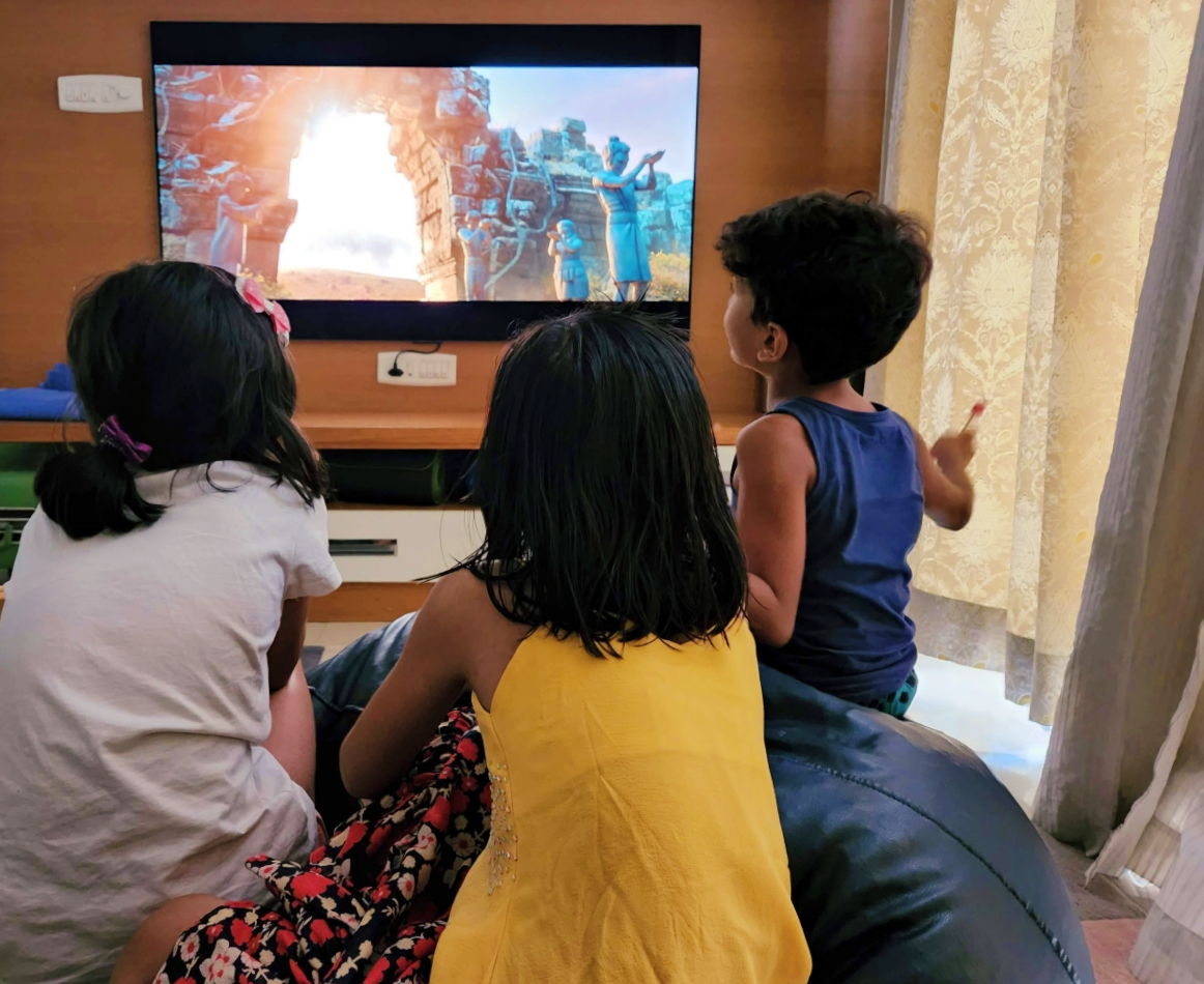 view of 3 kids from the back sitting on a beanbag watching a TV screen with a movie playing