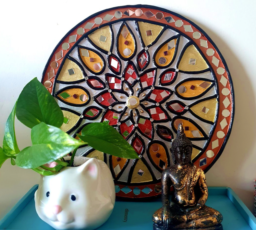 Mirror work and clay decorative piece in black and gold rests with a bronzed Buddha figurine and a small planter in the shape of a white cat with pothos plant growing