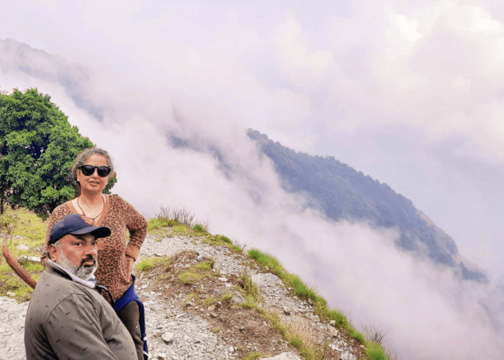 Man in cap sitting and woman standing next to him on a green hill surrounded by clouds on the mountains