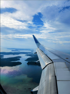view of the andaman and nicobar islands from the plane. the wing of the plane is jutting out and we can see small islands in a sea that's reflecting rainbow colours