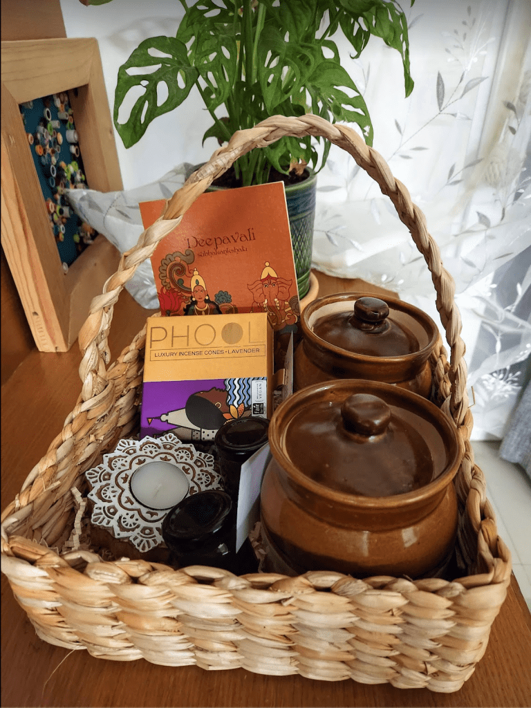 a reed basket with handle Diwali gift hamper with jars of traditional sweets, wooden block tea light, greeting card and box of Phool incense sticks