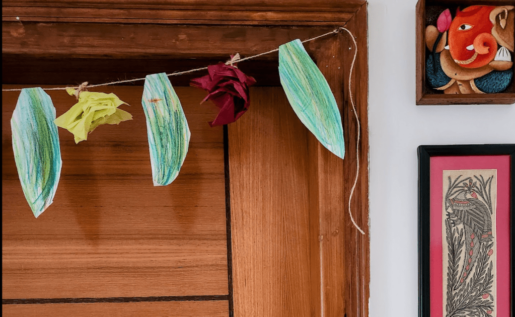 a string of green colored paper cut in a leaf shape with scrunched up paper flowers hung on the doorway. There's a painting of Ganesha and madhubani painting on the side wall