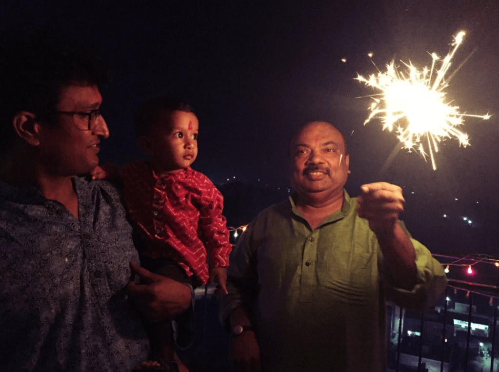 Father holding a toddler in his arms and both are looking at another man holding a lit up Diwali sparkler at night time on Diwali