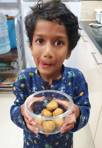 child in blue kurta holding a bowl of homemade Diwali sweets (Besan laddu) with his tongue licking his lips