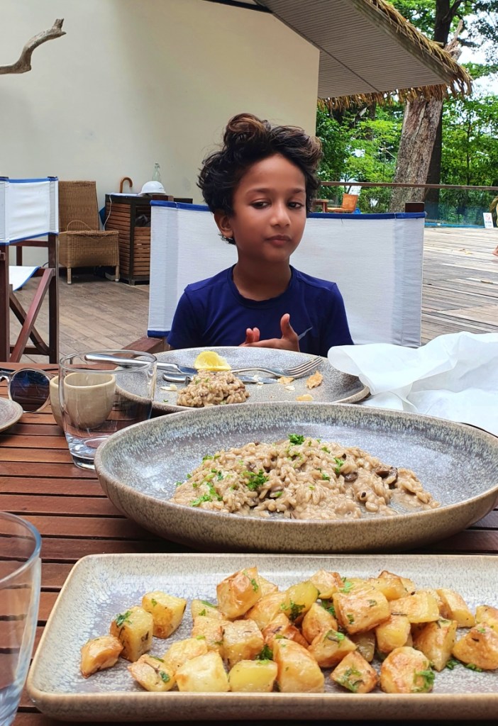 child in blue shirt sitting at a restaurant table looking at the food on the table which is a mushroom risotto and baked potatoes. restaurant at the taj exotica havelock islands