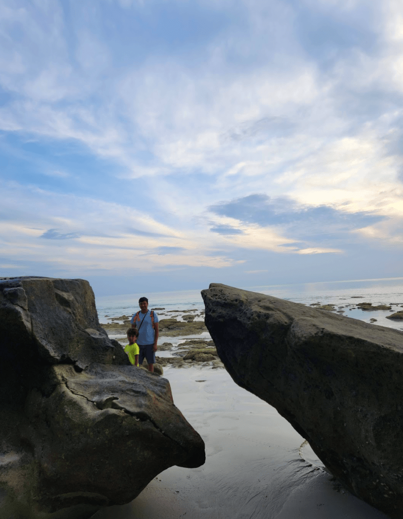 2 big black boulders on a beach with a narrow space between them where a man and his son are standing looking at the camera