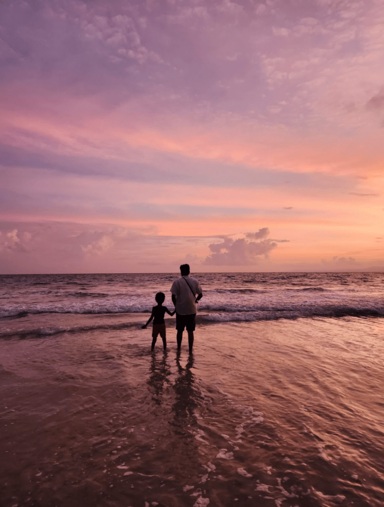 Father and son look towards the horizon from the beach as the waves roll in. The sky is different shades of purple as the sun sets on radhanagar beach on Havelock Island