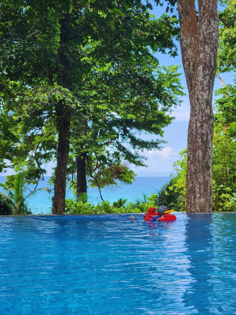 the infinity pool at the Taj Exotica Spa at Havelock island with the open sea in the background, and child in red floatie swimming in the pool under towering trees