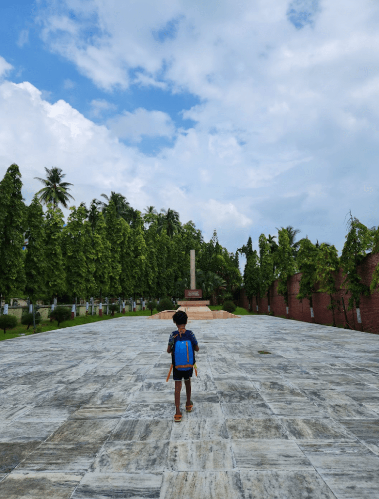 boy with a backpack walking towards a pillar that's a memorial to freedom fighters of India. Trees and blue skies line the path. This is in the Cellular Jail at Portt Blair