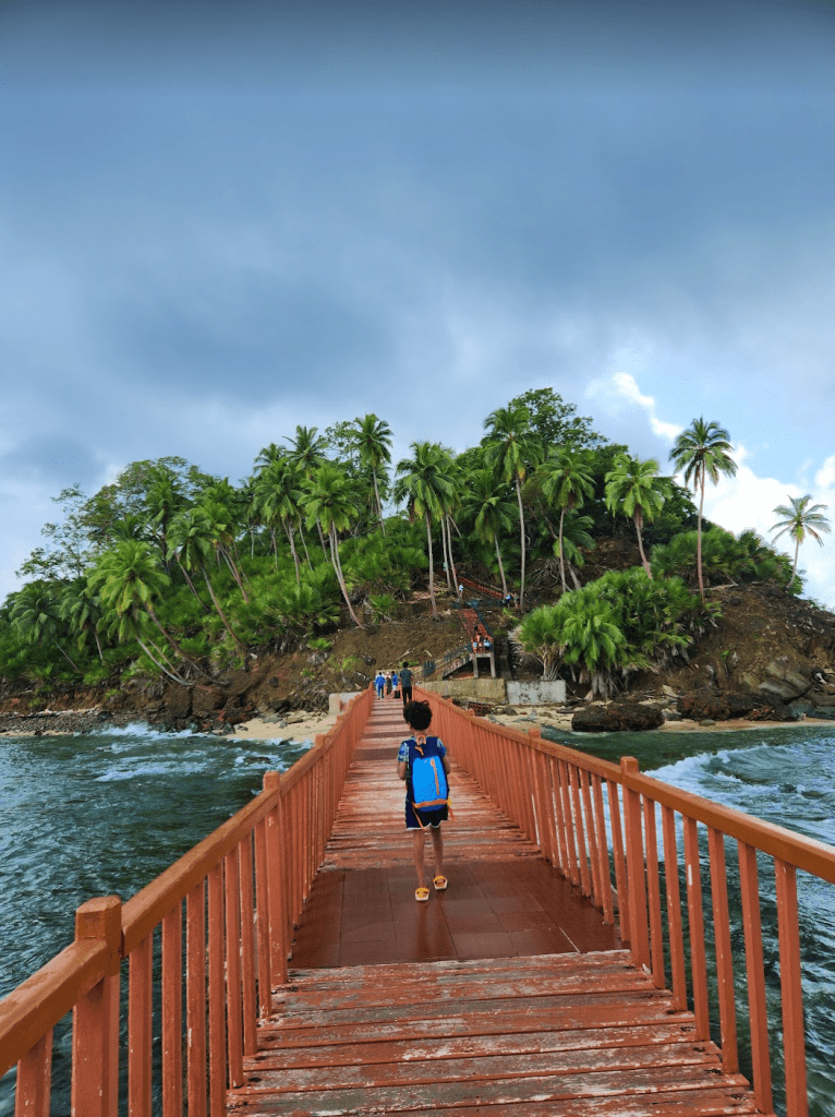 ross island as seen from the lighthouse with palm trees growing. A narrow wooden jetty connects the lighthouse to the island and a boy is walking across