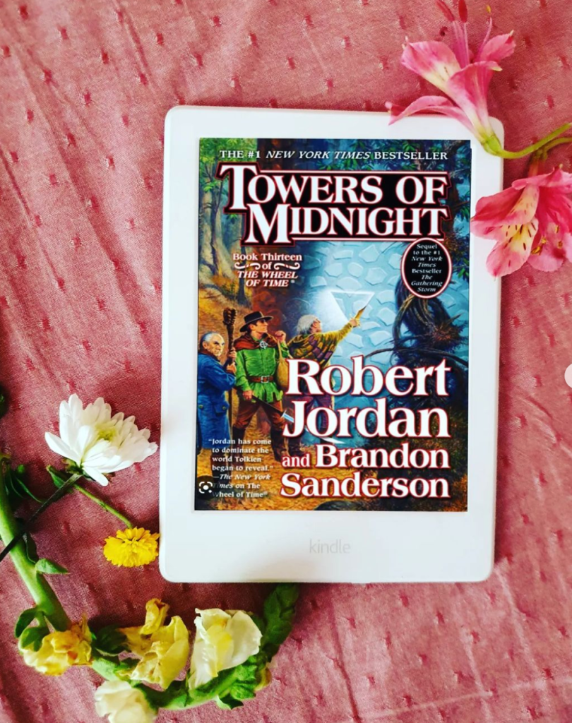 White Kindle with a colour cover of Towers of Midnight from the Wheel of Time series by Robert Jordan. The Kindle is on a pink sheet with flowers around it