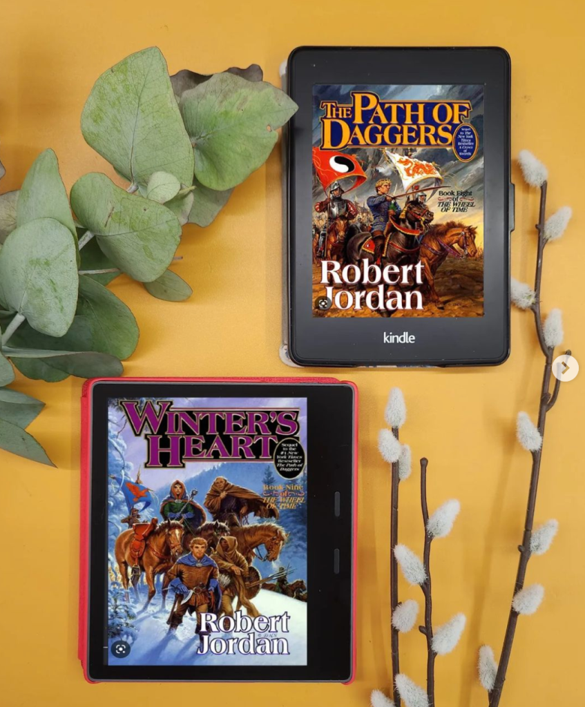 a Kindle Paperwhite and Kindle Oasis on a yellow background with leaves and sticks. The Kindles have the coloured covers of The Path of Daggers and Winters Heart from the Wheel of Time series b Robert Jordan