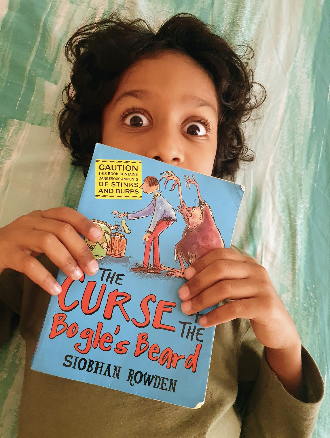 Child with wide eyes covering his lower face with the book by Siobhan Rowden named The Curse of Bogle's Beard