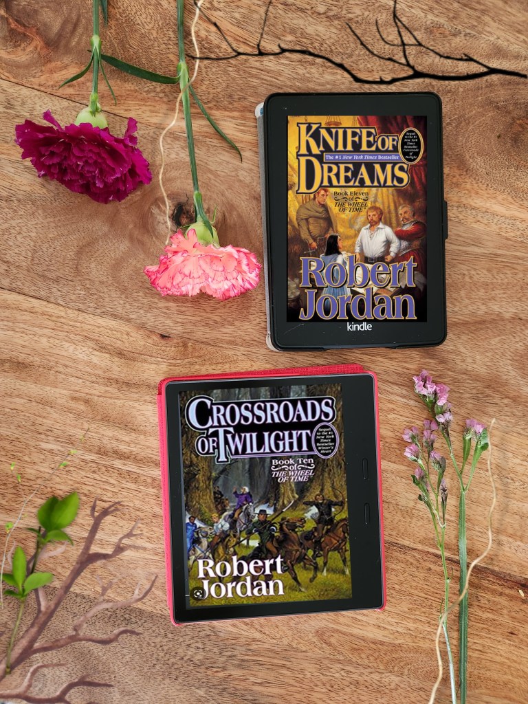 Robert Jordan's Wheel of Time Book 10 and 11 Crossroads of Twilight and Knife of Dreams on Kindle Oasis and Kindle Paperwhite on a wooden table with flower stems on the side