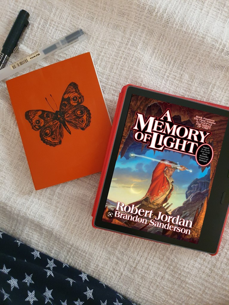 Robert Jordan's Wheel of Time Book 14 A Memory of Light cover on a Kindle Oasis with a notebook in red with a butterfly cover and 2 pens on a white bedcover