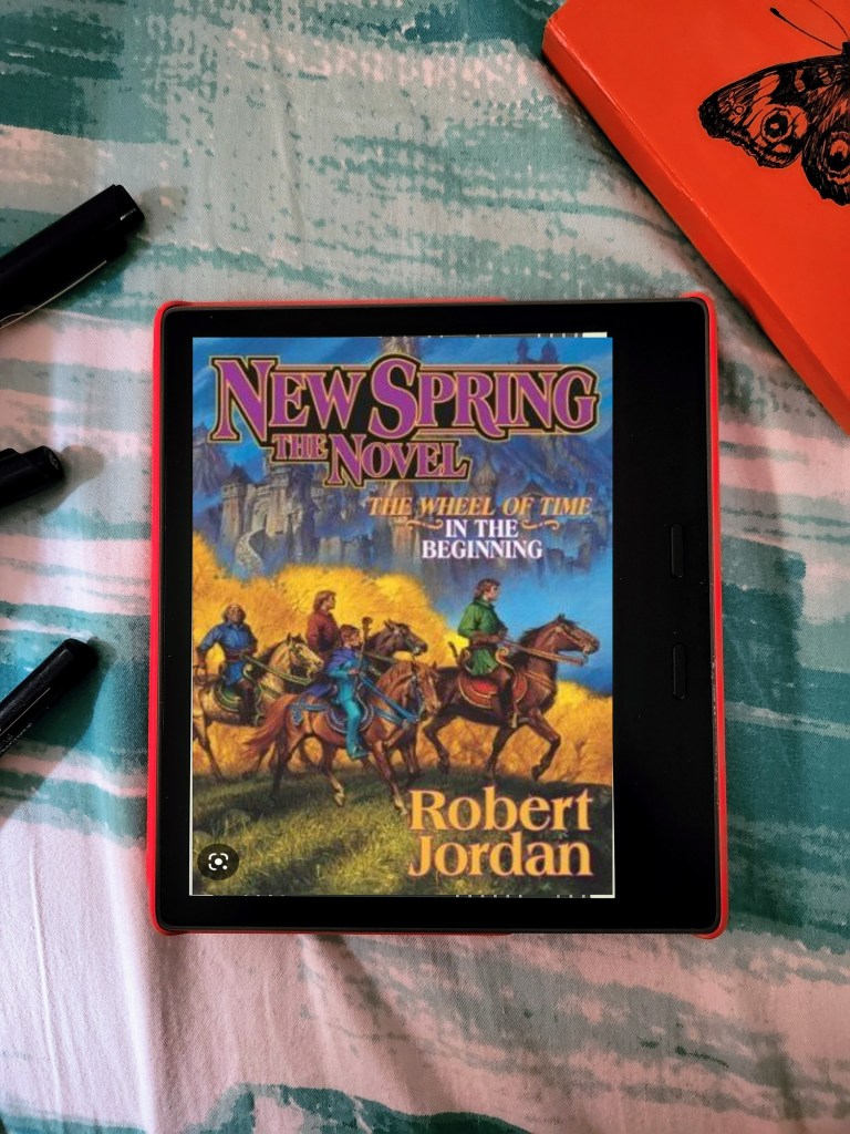 Cover of New Spring, A Wheel of Time prequel by Robert Jordan on a Kindle Oasis set on a green and white fabric with the edge of a red notebook and pens in the corner