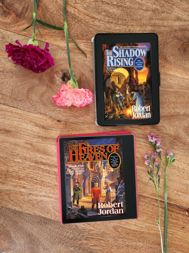 Robert Jordan's Wheel of Time Book 4 and 5 The Rising Shadow and The Fires of Heaven on Kindle Paperwhite and Kindle Oasis on a wooden table with flower stems on the side