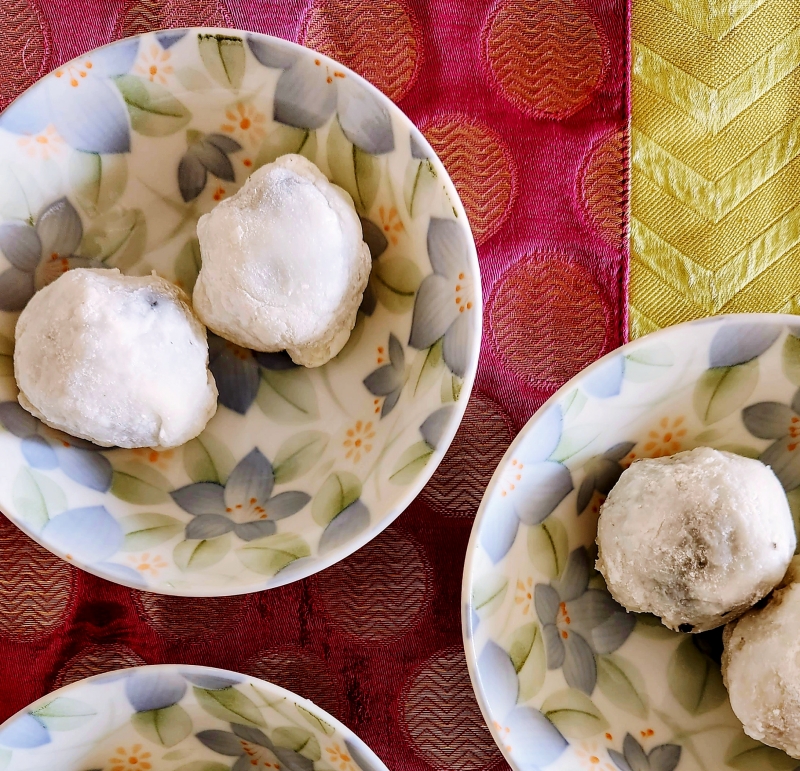 3 floral plates set on a vibrant pink and green mat. Each plate has 2 white mochi balls. One of the plate has the mochi balls split in half revealing the fillings - one with yellow mung dal and the other with black sesame seeds fillings