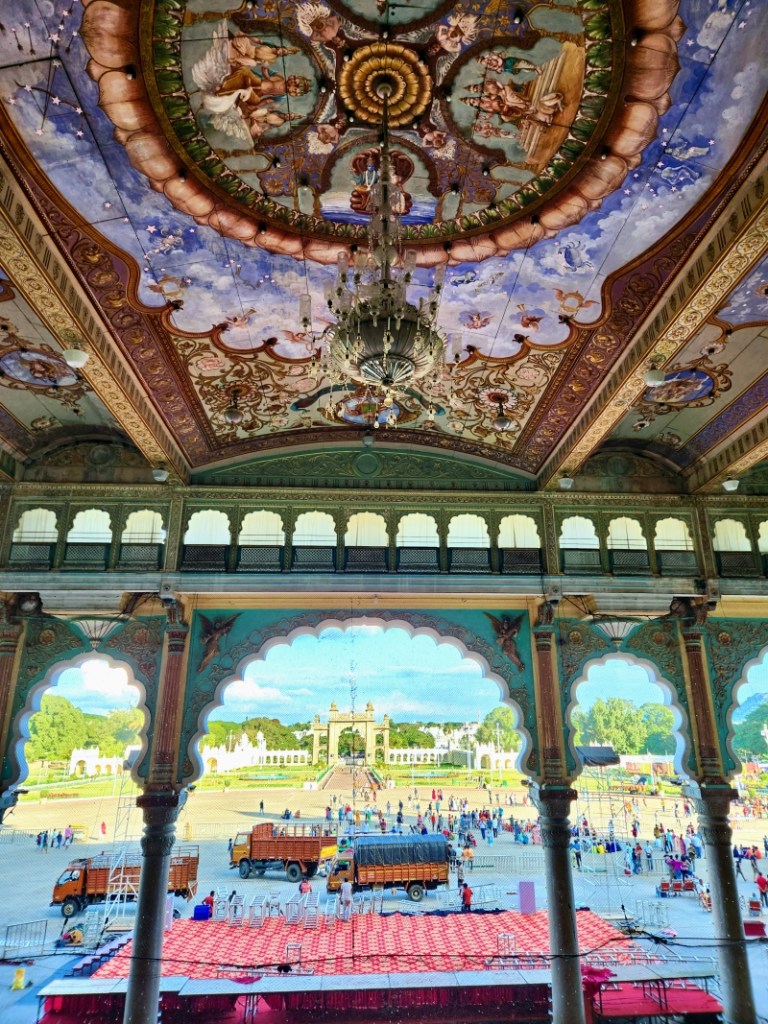 beautiful and ornately painted ceiling of the mysore palace with the arches on the facade showing the palace lawns and gate