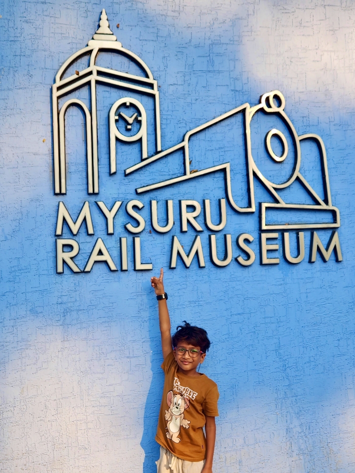boy in a brown tee shirt smiling and pointing to a sign that says Mysuru Rail museum